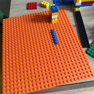 Puede incluir: Una placa base Lego naranja con varios ladrillos Lego de diferentes colores. Un ladrillo Lego azul está colocado en la placa base. Otras estructuras de Lego son parcialmente visibles en el fondo, lo que sugiere un tema de construcción.