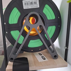 Puede incluir: Una bobina de filamento verde PLA Mate para impresi&oacute;n 3D, sujeta por un soporte met&aacute;lico gris. La bobina negra tiene una etiqueta que indica un peso de 1,1 kg. El soporte est&aacute; sobre una estanter&iacute;a de madera.