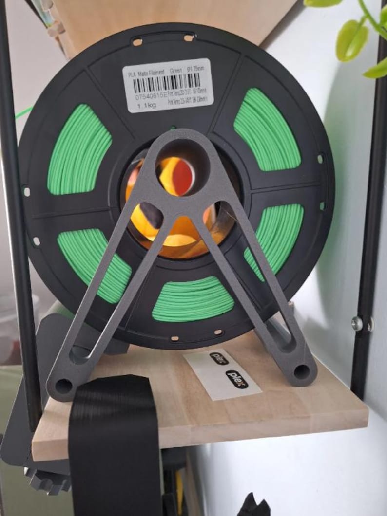 Puede incluir: Una bobina de filamento verde PLA Mate para impresi&oacute;n 3D, sujeta por un soporte met&aacute;lico gris. La bobina negra tiene una etiqueta que indica un peso de 1,1 kg. El soporte est&aacute; sobre una estanter&iacute;a de madera.