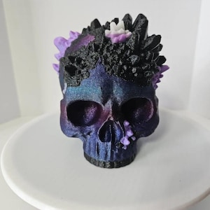 May include: A decorative skull sculpture with a crystal crown. The skull is painted in shades of blue, purple, and black with a metallic sheen. The crown features black and purple crystal formations, and small white accents.