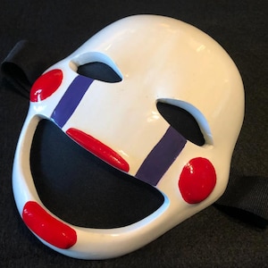 May include: A white mask with red and blue accents, featuring a smiling mouth and eye cutouts. The mask has red circles on the cheeks and a black strap. A decorative item.