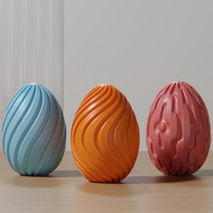 Könnte beinhalten: Drei dekorative eiförmige Objekte in Blau, Orange und Rosa. Das blaue Ei hat ein Spiraldesign, das orangefarbene Ei ein wellenförmiges Design und das rosa Ei ein strukturiertes Muster. Sie werden auf einer hellen Holzoberfläche präsentiert.