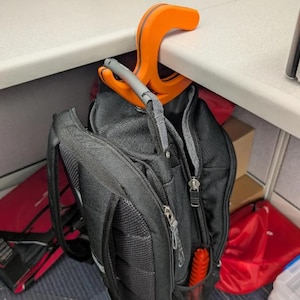 May include: An orange and gray backpack hook is attached to a white desk. A black backpack hangs from the hook. The hook is designed to hold a backpack or other items, keeping them off the floor.