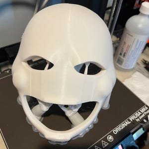 以下が含まれることがあります： 目と口の開口部がある白い3Dプリントマスク。マスクは黒い表面にあり、背景にはボトルとツールがあります。マスクは小道具またはより大きなプロジェクトの一部であるようです。