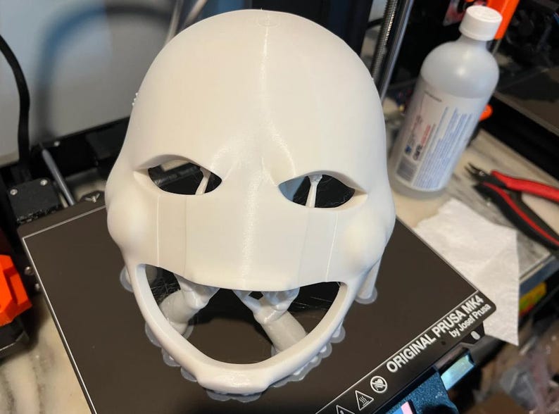 以下が含まれることがあります： 目と口の開口部がある白い3Dプリントマスク。マスクは黒い表面にあり、背景にはボトルとツールがあります。マスクは小道具またはより大きなプロジェクトの一部であるようです。