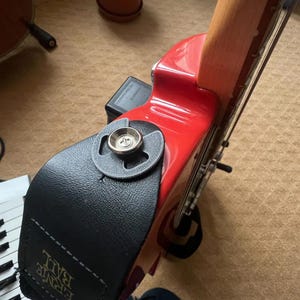 Puede incluir: Una guitarra eléctrica roja con una correa de cuero negro adjunta. La correa se asegura con un botón de metal. El mástil de la guitarra es de color marrón claro. La correa tiene el texto "Ernie Ball" impreso.