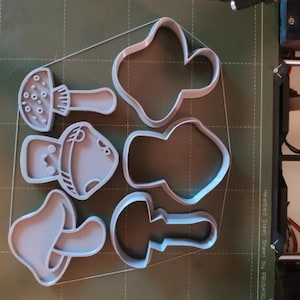 May include: A collection of gray mushroom-shaped cookie cutters in various designs, including a classic mushroom, a rounded mushroom, and a more abstract shape. The cutters are arranged on a green surface, likely a 3D printer bed.