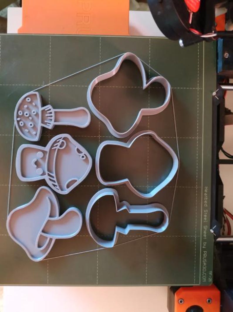May include: A collection of gray mushroom-shaped cookie cutters in various designs, including a classic mushroom, a rounded mushroom, and a more abstract shape. The cutters are arranged on a green surface, likely a 3D printer bed.