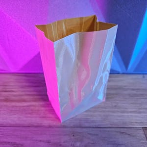 May include: A small, open-top paper bag with a bright pink exterior and a pearlescent, iridescent interior. The bag is sitting on a wooden surface, with a pink and blue background.