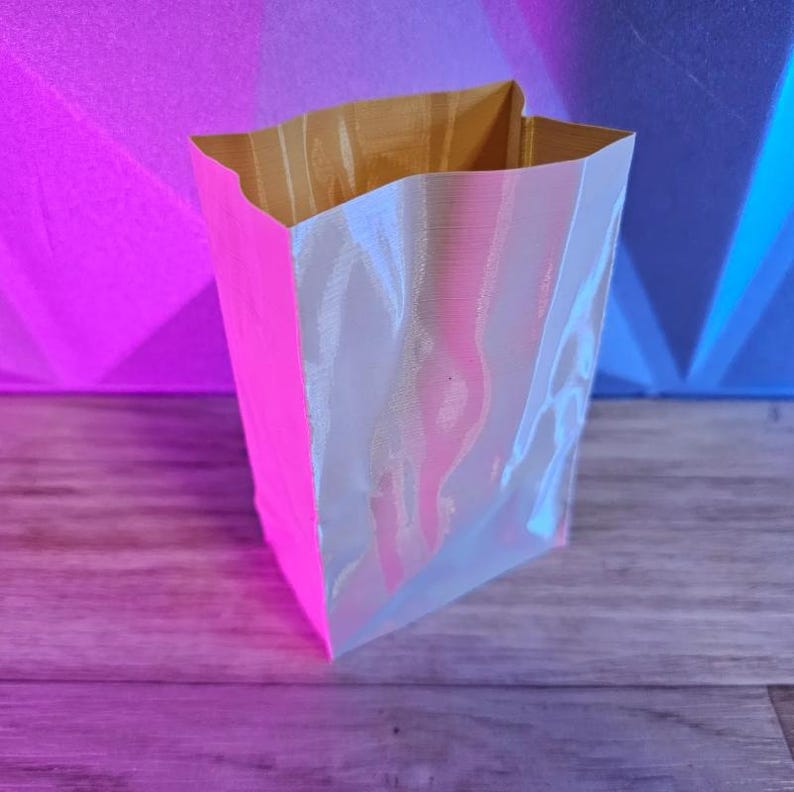 May include: A small, open-top paper bag with a bright pink exterior and a pearlescent, iridescent interior. The bag is sitting on a wooden surface, with a pink and blue background.