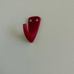 May include: A red metal wall hook with a curved design. The hook is mounted on a white wall and secured with two small screws. The hook is approximately 3 inches in height.