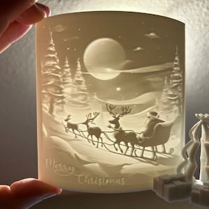 Könnte beinhalten: Ein weißer weihnachtlicher Kerzenhalter mit einer Winterlandschaft, die den Weihnachtsmann in einem von Rentieren gezogenen Schlitten, einen Vollmond und die Worte "Merry Christmas" zeigt. Enthält einen kleinen Weihnachtsbaum und zwei Geschenkboxen.