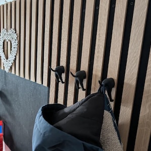 May include: A wooden wall panel with vertical wood slats and black accents. Three black dog-shaped hooks are mounted on the panel, with a blue jacket hanging from one. Decorative items, including a heart-shaped ornament, are also visible.