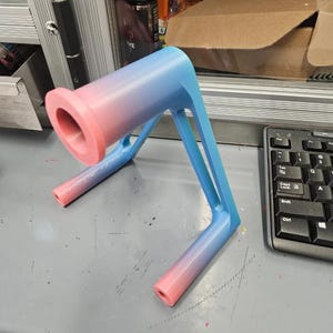 Puede incluir: Objeto impreso en 3D con un degradado rosa y azul. Presenta una parte superior cil&iacute;ndrica y patas anguladas para soporte. El color cambia de rosa a azul a lo largo de su longitud. Parece ser un soporte.