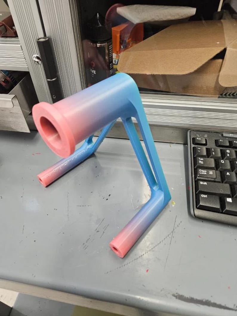 Puede incluir: Objeto impreso en 3D con un degradado rosa y azul. Presenta una parte superior cil&iacute;ndrica y patas anguladas para soporte. El color cambia de rosa a azul a lo largo de su longitud. Parece ser un soporte.