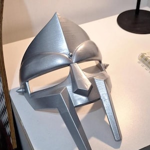 Puede incluir: Una máscara plateada con un diseño angular distintivo. La máscara presenta una parte superior puntiaguda, una placa facial central y dos secciones inferiores con detalles afilados. La máscara está hecha de un material metálico y es probablemente una pieza de disfraz.