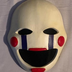 以下が含まれることがあります： 黒い目の穴、赤い頬、赤い口が付いた白いマスク。目の下に青と紫の涙の筋があります。マスクは小道具またはコスチュームの一部です。
