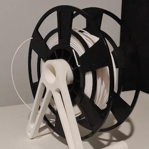 Puede incluir: Una bobina de filamento para impresora 3D en blanco y negro con un rollo de filamento blanco. La bobina est&aacute; sujeta por un soporte blanco. El soporte tiene una base curva y soportes angulados. El filamento es una l&iacute;nea blanca continua.