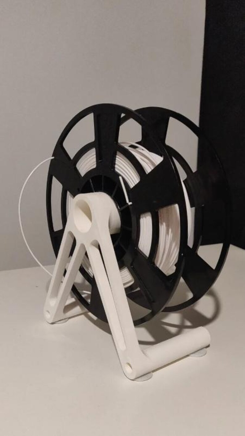 Puede incluir: Una bobina de filamento para impresora 3D en blanco y negro con un rollo de filamento blanco. La bobina est&aacute; sujeta por un soporte blanco. El soporte tiene una base curva y soportes angulados. El filamento es una l&iacute;nea blanca continua.