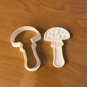 May include: Two white plastic mushroom-shaped cookie cutters. One cutter is a complete mushroom shape, and the other is an outline of a mushroom. The cutters are on a wooden surface.