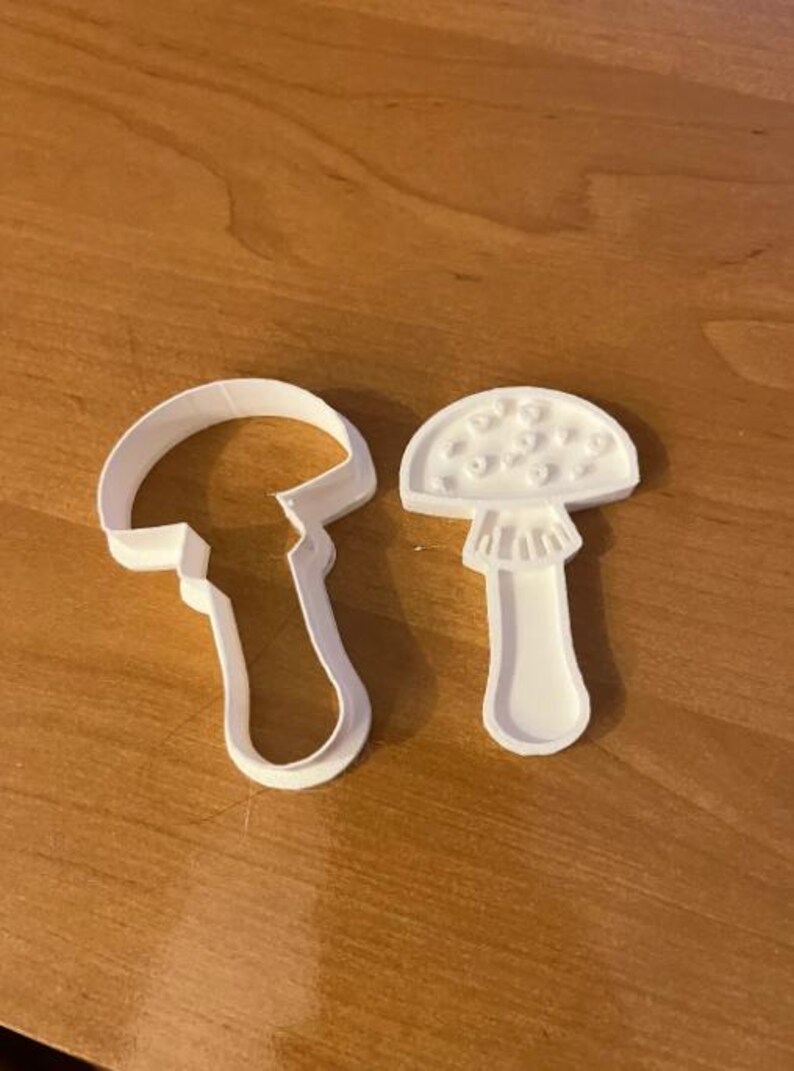 May include: Two white plastic mushroom-shaped cookie cutters. One cutter is a complete mushroom shape, and the other is an outline of a mushroom. The cutters are on a wooden surface.