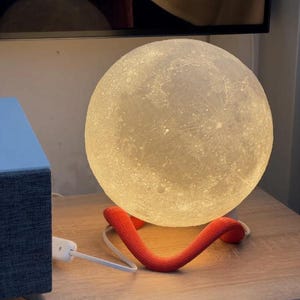 May include: A spherical, illuminated moon lamp with a textured surface, resting on a red, curved support. The lamp is plugged into a power outlet with a white cord. The lamp emits a soft, warm glow.