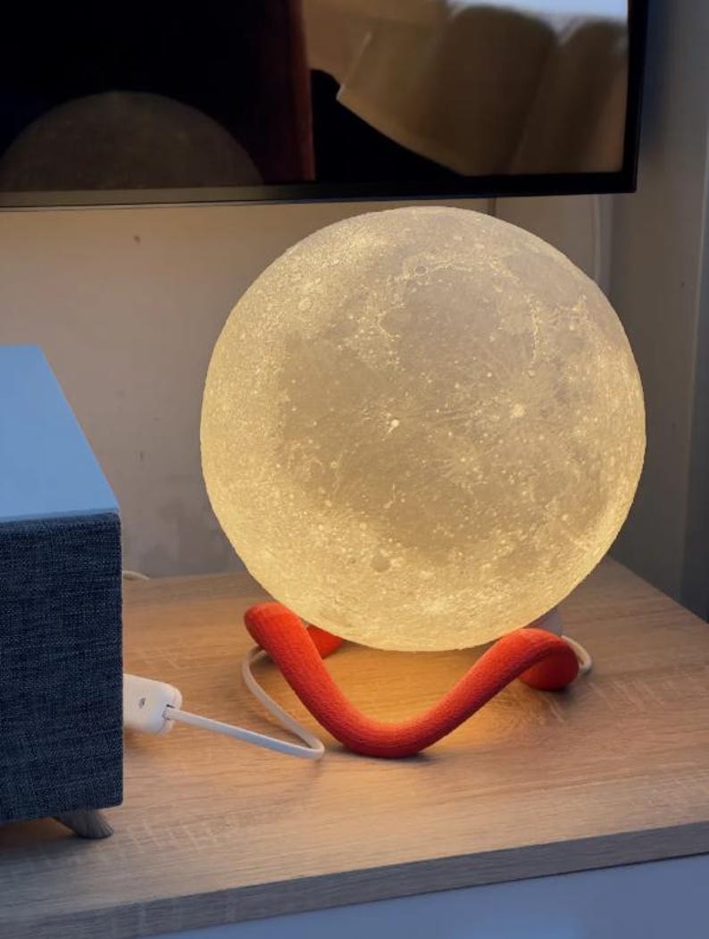 May include: A spherical, illuminated moon lamp with a textured surface, resting on a red, curved support. The lamp is plugged into a power outlet with a white cord. The lamp emits a soft, warm glow.