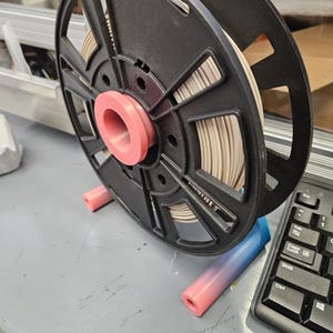 Puede incluir: Una bobina de filamento de impresora 3D negra con centro rosa y filamento beige. La bobina est&aacute; montada sobre un soporte rosa y azul, junto a un teclado de ordenador. La configuraci&oacute;n est&aacute; sobre una superficie gris.