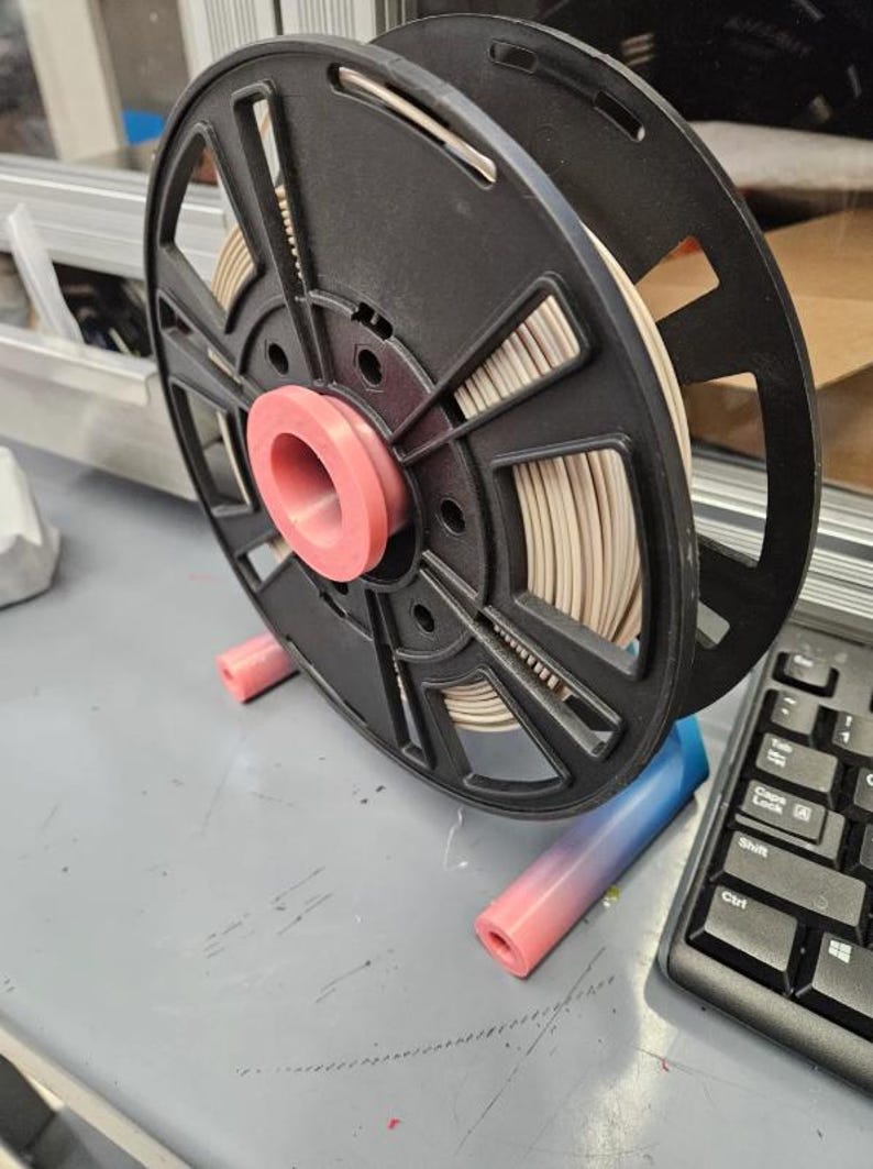 Puede incluir: Una bobina de filamento de impresora 3D negra con centro rosa y filamento beige. La bobina est&aacute; montada sobre un soporte rosa y azul, junto a un teclado de ordenador. La configuraci&oacute;n est&aacute; sobre una superficie gris.