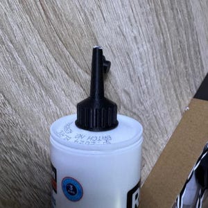 May include: A white bottle with a black nozzle and cap. The bottle has a blue and red label with text and a blue circular sticker. The top of the bottle has the text "BATCH NO" and other text.