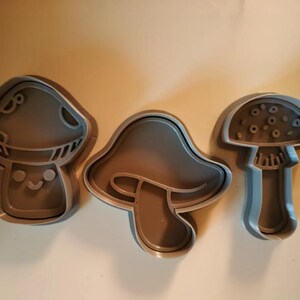 May include: Three gray mushroom-shaped cookie cutters. One has a smiling face, another has a simple outline, and the third has a detailed cap. These cutters are ideal for creating themed cookies.