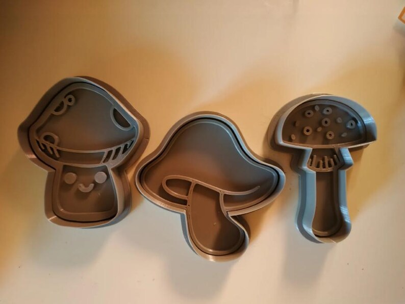 May include: Three gray mushroom-shaped cookie cutters. One has a smiling face, another has a simple outline, and the third has a detailed cap. These cutters are ideal for creating themed cookies.
