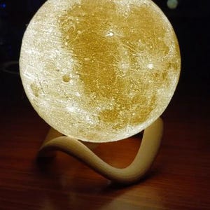 May include: A spherical, illuminated moon lamp with a textured surface, resting on a curved, light-colored wooden base. The lamp emits a warm, golden glow, creating a soft ambiance. The lamp is a decorative home decor item.