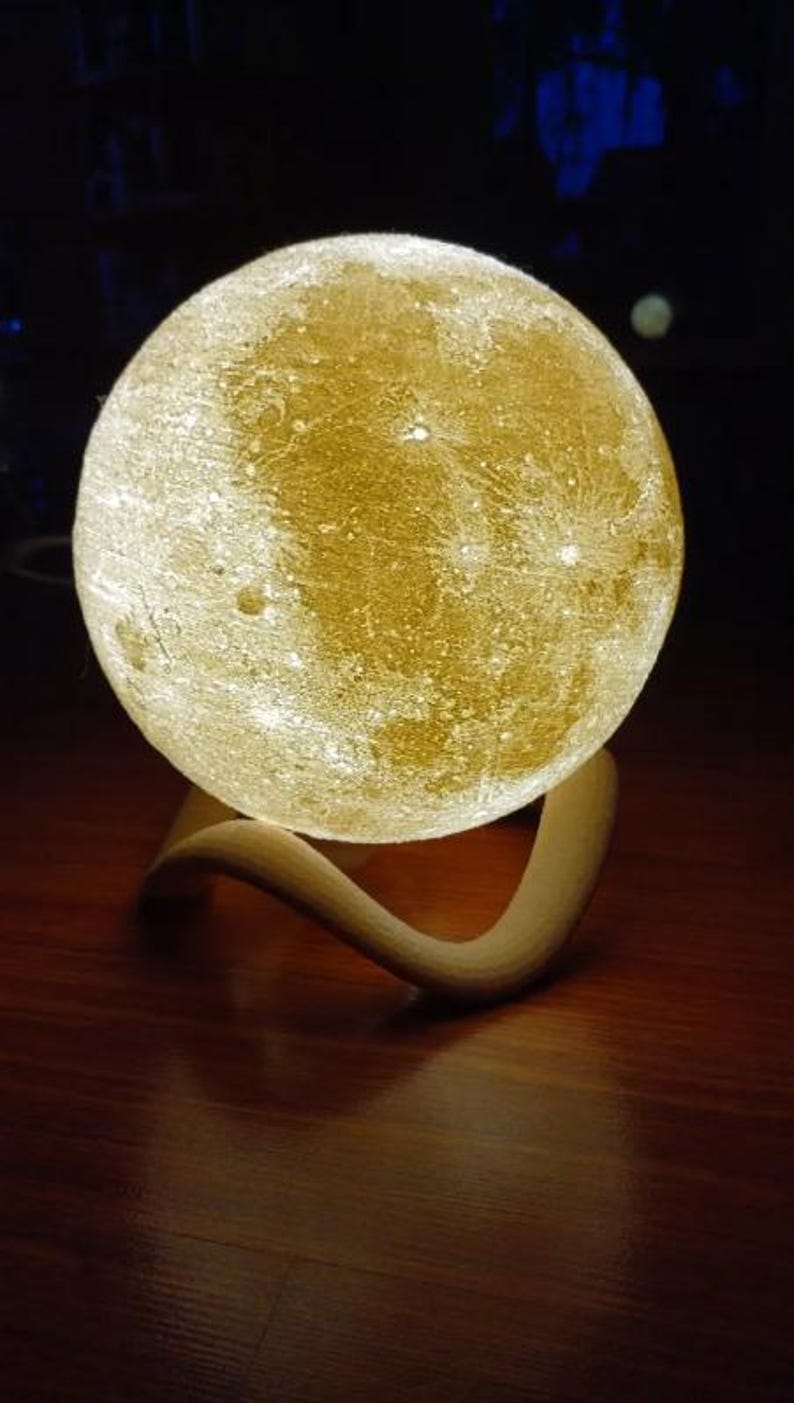 May include: A spherical, illuminated moon lamp with a textured surface, resting on a curved, light-colored wooden base. The lamp emits a warm, golden glow, creating a soft ambiance. The lamp is a decorative home decor item.