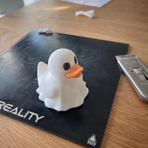 May include: A 3D-printed white duck figurine with a rounded body, small black eyes, and an orange beak. The duck is sitting on a black surface with the word "REALITY" visible. A utility knife is on the right side of the image.
