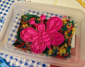 May include: A clear plastic bin filled with colorful pasta and a bright pink butterfly-shaped toy. The pasta includes green, red, and yellow varieties, creating a visually appealing sensory experience. The butterfly toy is centered in the bin.