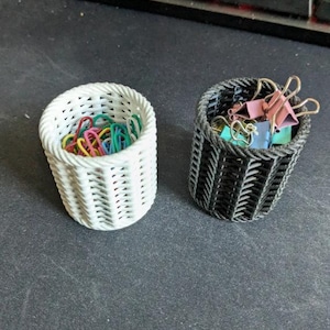 May include: Two small, woven baskets. One is white and filled with colorful paper clips. The other is black and contains binder clips. The baskets are cylindrical with a textured, open weave design.