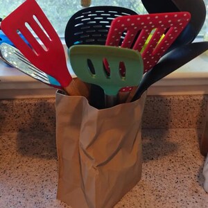May include: A collection of kitchen utensils, including spatulas, slotted spoons, and a ladle, are stored in a brown paper bag. The utensils are various colors, including red, green, and black. The bag sits on a speckled countertop.