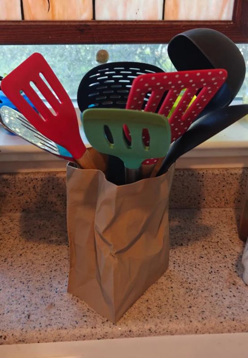 May include: A collection of kitchen utensils, including spatulas, slotted spoons, and a ladle, are stored in a brown paper bag. The utensils are various colors, including red, green, and black. The bag sits on a speckled countertop.