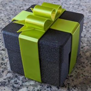 Może przedstawiać: Czarny pudełko na prezent o teksturowanej powierzchni, ozdobione jasnozieloną wstążką i kokardą. Pudełko jest kwadratowe, a wstążka owija się wokół niego, tworząc świąteczną prezentację. Kokarda jest wyśrodkowana na górze.