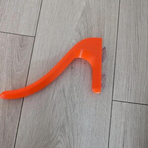 May include: Bright orange, curved plastic object with a handle-like design. It has two clear suction cups attached to one end. The object is resting on a light-colored wooden surface.