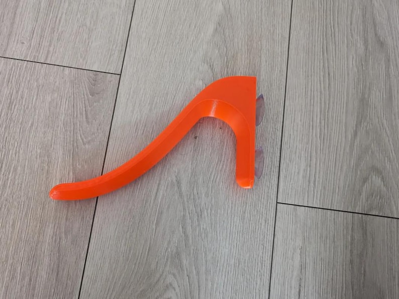 May include: Bright orange, curved plastic object with a handle-like design. It has two clear suction cups attached to one end. The object is resting on a light-colored wooden surface.