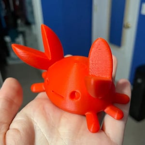 May include: A bright orange, 3D-printed crab figurine. The crab has a rounded body, six legs, and two large, upward-pointing claws. The figurine is held in a hand, with a blue background visible.