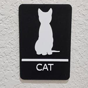 Pode incluir: Placa retangular preta com uma silhueta branca de um gato sentado. A palavra "CAT" é impressa em branco abaixo da imagem do gato, acima de uma linha horizontal branca.