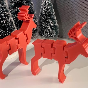 Pode incluir: Duas figuras de renas articuladas vermelhas. As renas são feitas de blocos conectados, permitindo o movimento. As hastes são detalhadas e as pernas são posicionadas para ficar em pé. O fundo inclui árvores de Natal em miniatura.