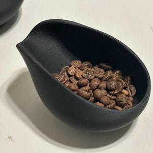 May include: A black, scoop-shaped container filled with roasted coffee beans. The container has a smooth, matte finish and a curved design. The coffee beans are various shades of brown and tan, with visible textures.