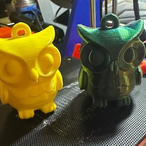 May include: Two 3D-printed owl figurines. One is bright yellow, the other is a dark green with a metallic sheen. Both have large eyes, a small beak, and a loop at the top for hanging. They are sitting on a black surface.