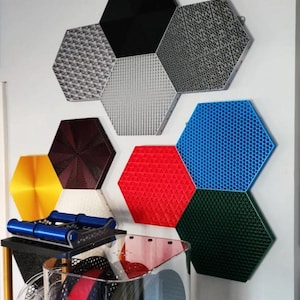 May include: A collection of hexagon-shaped wall panels in various colors and textures. The panels are mounted on a white wall and include black, gray, red, blue, green, and yellow options. Some panels have textured surfaces, while others are smooth.