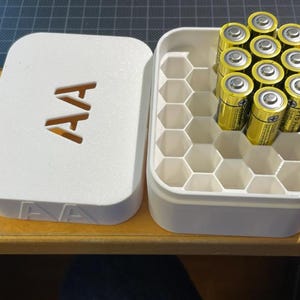 May include: A white plastic battery organizer with a lid, featuring the letters "AA" cut out. The open container holds several gold and black AA batteries. The interior of the container is divided into hexagonal compartments.