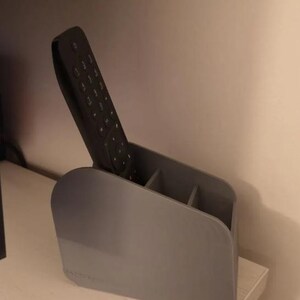 May include: A grey plastic remote control tidy with three compartments, holding a black remote. The tidy is on a white surface, with a light-coloured wall in the background. The holder is designed to organise remotes.